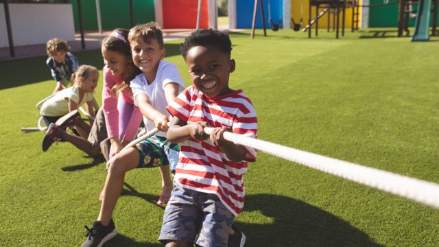 Front view of multi ethnic group of happy school kids playing tug of war in playground