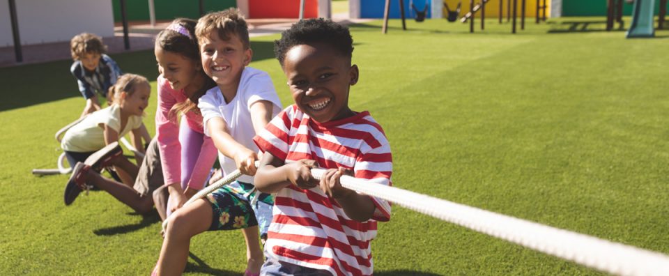 Front view of multi ethnic group of happy school kids playing tug of war in playground